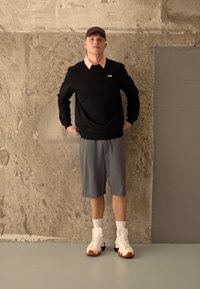 Felpa nera con colletto rosa, pantaloni corti grigi e sneakers bianche con dettagli arancioni, in piedi davanti a uno sfondo in cemento testurizzato.