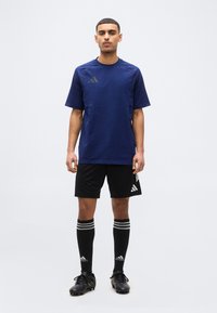 Άνδρας που στέκεται με μπλε αθλητική μπλούζα, μαύρο σορτς, μαύρες κάλτσες μέχρι το γόνατο με λευκές ρίγες και μαύρα ποδοσφαιρικά παπούτσια, σε λευκό φόντο.