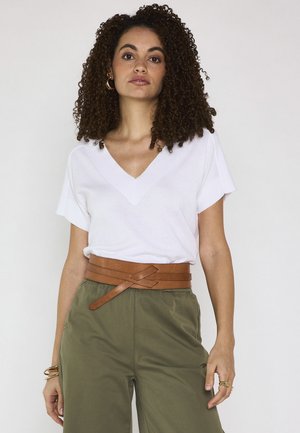 Femme aux cheveux bouclés portant un t-shirt blanc à col en V, un pantalon vert olive et une ceinture marron nouée sur un fond uni.