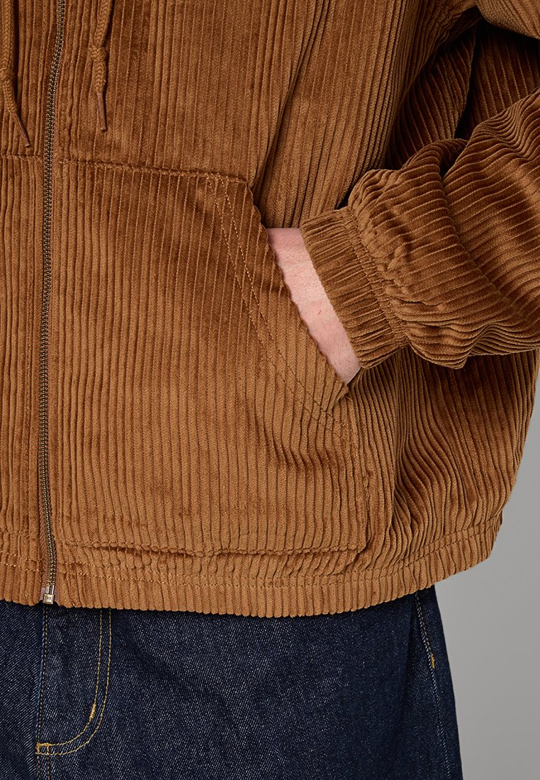 Primer plano de una mano en un bolsillo de una chaqueta de pana marrón combinada con jeans de denim azul oscuro.