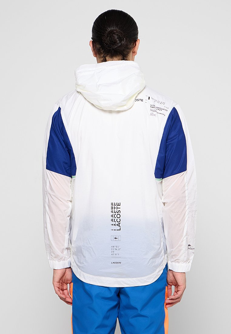 Veste blanche légère avec des accents bleus, une capuche et des détails imprimés. Présente un effet dégradé et des manches côtelées pour un ajustement confortable.