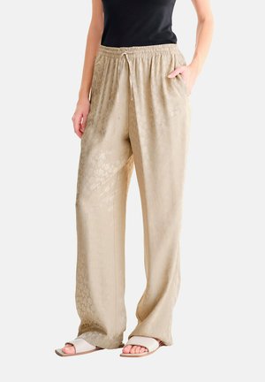 Persoon die beige wijde broek met subtiel patroon draagt, een taille met trekkoord, een zwarte top en witte open sandalen, staand met één hand in de zak.