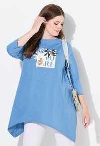 Himmelblaues, langärmliges Tunika mit asymmetrischem Saum, versehen mit einem grafischen Design in Creme- und dunklen Farben. Kombiniert mit weißen Leggings.
