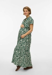 Femme enceinte aux cheveux courts portant une robe maxi verte et blanche à motifs et des chaussures marron, tenant doucement son ventre et souriant.