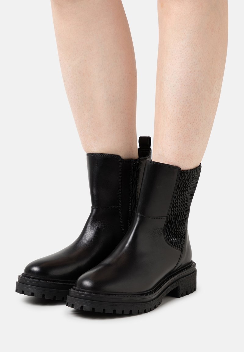 Bottes basses en cuir noir avec un panneau en maille texturée, design imperméable, semelle en caoutchouc épaisse et fermeture éclair sur le côté.