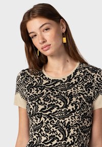 Camiseta de punto de manga corta en negro y crema con un patrón floral, escote redondo y textura ligera; modelo lleva pendientes de bloque dorados.
