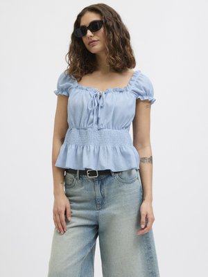 Light blue peplum top with ruffled sleeves, featuring a tie closure at the front, paired with light-washed wide-leg jeans. Black sunglasses worn.