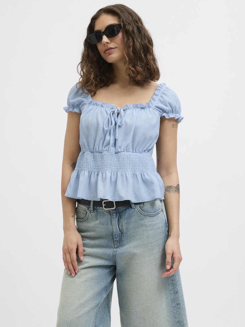 Light blue peplum top with ruffled sleeves, featuring a tie closure at the front, paired with light-washed wide-leg jeans. Black sunglasses worn.