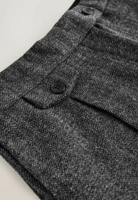 Close-up of gray herringbone fabric trousers with a black button on the waistband and a side pocket flap.