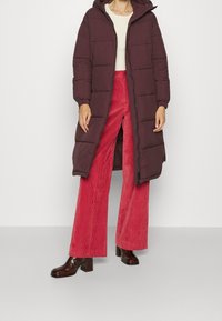 Langer bordeauxfarbener Puffer-Mantel mit Kapuze, genähten Paneelen, getragen über einem cremefarbenen Oberteil und roten, weiten Cordhosen; glänzende braune Blockabsatzstiefel.