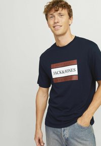 Mörkblå t-shirt i bomullsmaterial med en rektangulär logotyp med "JACK&JONES" i vitt och rött på framsidan. Korta ärmar, rundad hals.
