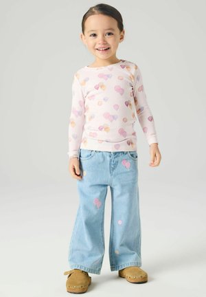 Nasmejana mlada deklica s temnimi lasmi v svetli majici z dolgimi rokavi, obdana s pastelnimi baloni, v svetlo modrih širokih kavbojkah in rjavih natikačih.