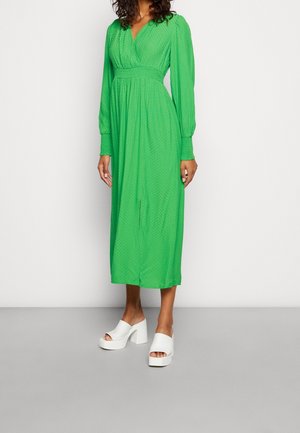 Długa zielona sukienka z długim rękawem, dekoltem w serek, talią podkreśloną paskiem oraz zwiewną spódnicą. Wykonana z fakturowanej tkaniny, z rozcięciem z przodu. Kompletuje ją białe sandały na platformie.