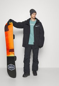 Snowboardeur portant une veste et un pantalon isolés noirs, un bonnet gris et des gants, tenant un snowboard orange et noir avec un logo.