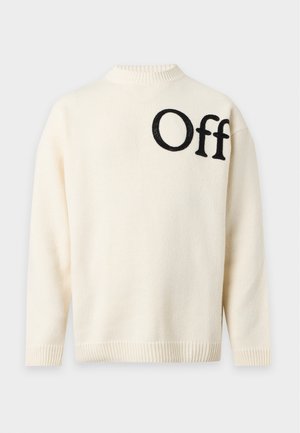 Jeune homme debout portant un pull crème avec le texte ""Off"" sur la poitrine, un pantalon de survêtement gris clair et des baskets blanches sur fond blanc.