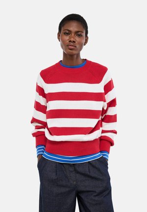 Jeune personne portant un pull rayé rouge et blanc par-dessus une chemise rayée bleu et blanc avec un pantalon en jean foncé, debout les mains dans les poches.