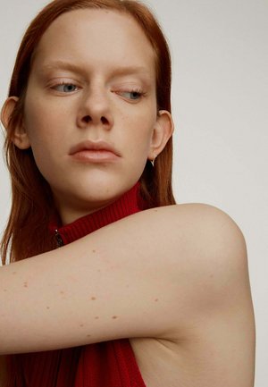 Prenda roja de cuello alto con textura acanalada, que presenta un cierre. Piel clara con pecas, mostrando un brazo descubierto. El fondo es de tonos neutros, lo que realza el enfoque en el sujeto.