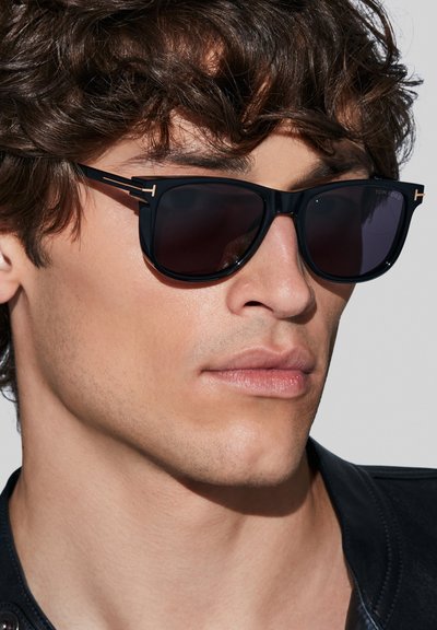 Occhiali da sole neri con lenti scure presentano un design elegante, dettagli dorati e una finitura opaca, incorniciati da una forma rettangolare.