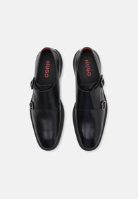 Zwarte leren schoenen met een afgeronde neus, voorzien van een zijgesp en een gladde textuur. Binnenin staat "HUGO" in rode print.