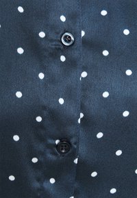 Camicia button-up navy in tessuto liscio con pois bianchi. Presenta un bottone nero al colletto e una leggera lucentezza.
