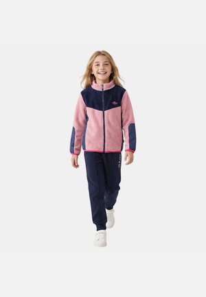 Enfant souriant marchant vers l'avant portant une veste polaire zippée rose et marine, un pantalon de survêtement marine et des baskets blanches sur fond uni.