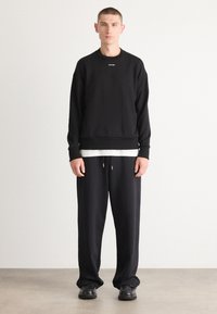 Schwarzer Pullover mit Rundhalsausschnitt, mit kleinem Logo auf der Vorderseite; kombiniert mit lockeren schwarzen Jogginghosen und schwarzen Schuhen.