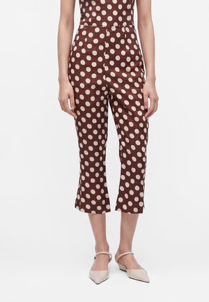 Kvinde iført brune korte bukser med store beige polkaprikker og beige spidse ballerinaer med ankelstropper, stående mod en hvid baggrund.