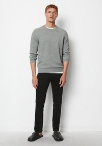 Mann steht an einer weißen Wand und trägt einen grauen Pullover, schwarze Hosen und schwarze Slipper.