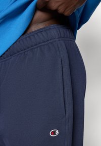 Μπλε παντελόνι φόρμας με ελαστική μέση, πλαϊνές τσέπες και μικρό κε ντημένο λογότυπο σε κόκκινο και λευκό στο κάτω μέρος του ποδιού. Μαλακή υφή υφάσματος.