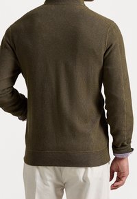 Muž v olivově zeleném pleteném svetru s texturou a světlých béžových kalhotách, stojící s jednou rukou na bok, zobrazený zezadu.