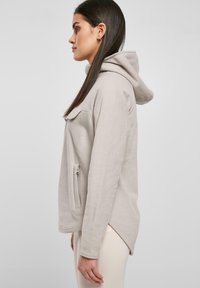 Lys grå fleece hoodie med en kængurulomme, lynlåsdetalje på siden og en afslappet pasform, der fremhæver en glat tekstur og hætte.