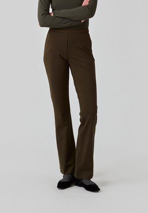 Person trägt eng anliegende dunkelolivgrüne Hose, graue Socken, schwarze Schuhe und ein passendes langärmeliges Oberteil mit verschränkten Armen.