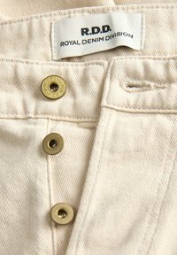 Gros plan d’un denim couleur crème avec trois boutons en bronze et une étiquette en tissu indiquant R.D.D. Royal Denim Division.