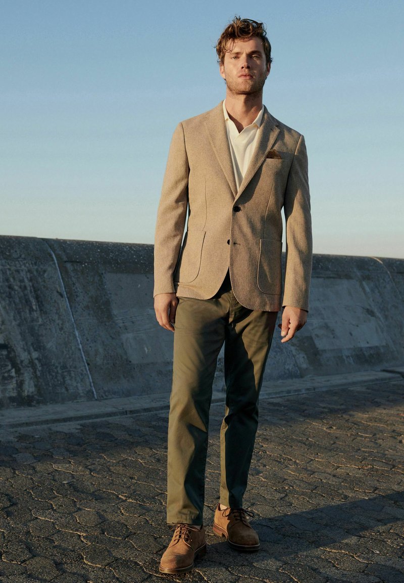 Blazer beige ajustado sobre una camisa blanca; pantalones verde oliva; zapatos de ante marrón. Diseño sencillo con bolsillos y un corte clásico.
