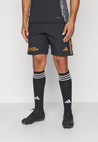 Schwarze Sportshorts mit goldener Branding, kombiniert mit knielangen schwarzen Socken mit weißen Streifen und schwarzen Fußballschuhen.