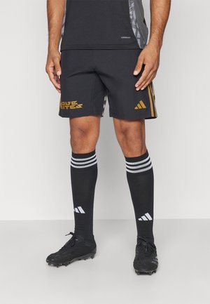 Zwarte sportshorts met gouden branding, gedragen met knielange zwarte sokken met witte strepen en zwarte voetbalschoenen.