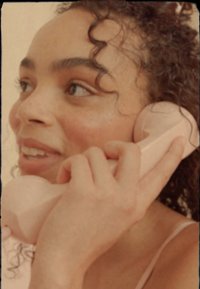 Rosa vintage-telefon hålls mot ansiktet. Personen har lockigt hår, en ljus topp och ler lätt, visar mjuk hud och minimal makeup.