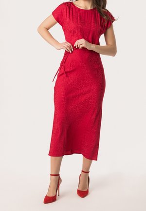 Kvinde iført en tætsiddende rød midi-kjole med korte ærmer og matchende røde højhælede sko med ankelrem, stående foran en ensfarvet baggrund.