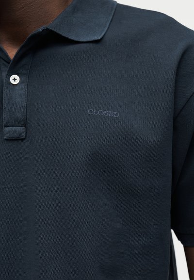 Πόλο μπλε σκούρο με λευκό κουμπί και διακριτικό κεντημένο κείμενο "CLOSED" στο στήθος, φορεμένο από άτομο με σκούρο τόνο δέρματος.