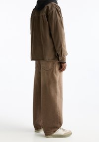 Man die met de rug naar de camera staat, gekleed in bruin wijde spijkerbroeken, een bijpassende bruine jas over een zwart hoodie en witte sneakers, tegen een witte achtergrond.