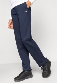 Σκούρο μπλε φόρμας από μαλακό ύφασμα με πλαϊνές τσέπες, διακριτικό λογότυπο στο ύψος του ισχίου και ίσια γραμμή στα μπατζάκια.