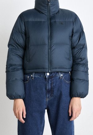 Persoon draagt een korte marineblauwe pufferjas met een ritssluiting aan de voorkant en blauwe jeans met een hoge taille, staand tegen een effen achtergrond.