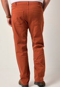 Man som bär rostfärgade byxor och bruna skor, stående med händerna bakom ryggen mot en enkel ljus bakgrund.