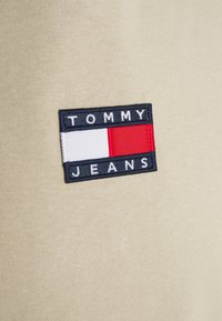 Tommy Jeans logo patch with white, red, and navy blue rectangles stitched on beige fabric.