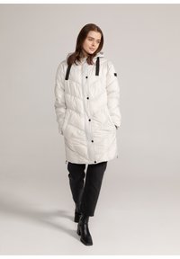 Zachte witte parka met een capuchon en zwarte accenten. Bevat een chevron-quiltpatroon, knopen aan de voorkant en zijzakken. Draagt over zwarte broeken.