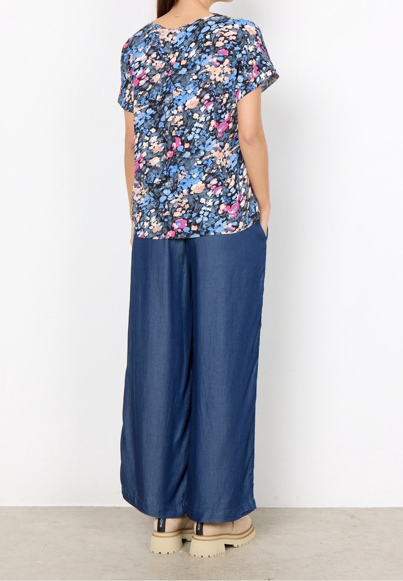 Femme portant un haut à manches courtes à fleurs bleu, rose et blanc, associé à un pantalon large bleu foncé et des chaussures épaisses beige, debout contre un mur blanc.