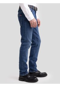 Férfi kék farmerben, fehér ingben, fekete övvel és fekete bőr cipőben, egyszerű világos háttér előtt áll.