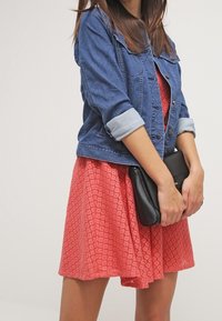 Veste en denim aux manches retroussées portée sur une robe corail à motifs, tenant une pochette noire. Mise en avant des textures et des couleurs des vêtements.