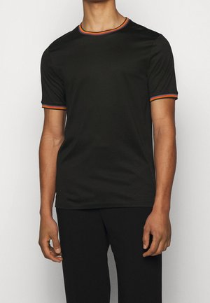 Uomo che indossa una maglietta nera a maniche corte con strisce rosse, arancioni e gialle sul colletto e sul bordo delle maniche, in piedi davanti a uno sfondo semplice.