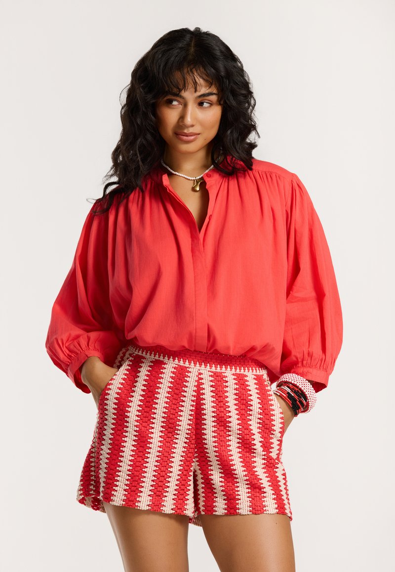 Blusa corallo a maniche lunghe con colletto bottone; abbinata a pantaloni corti intrecciati a strisce rosse e bianche con un motivo strutturato.